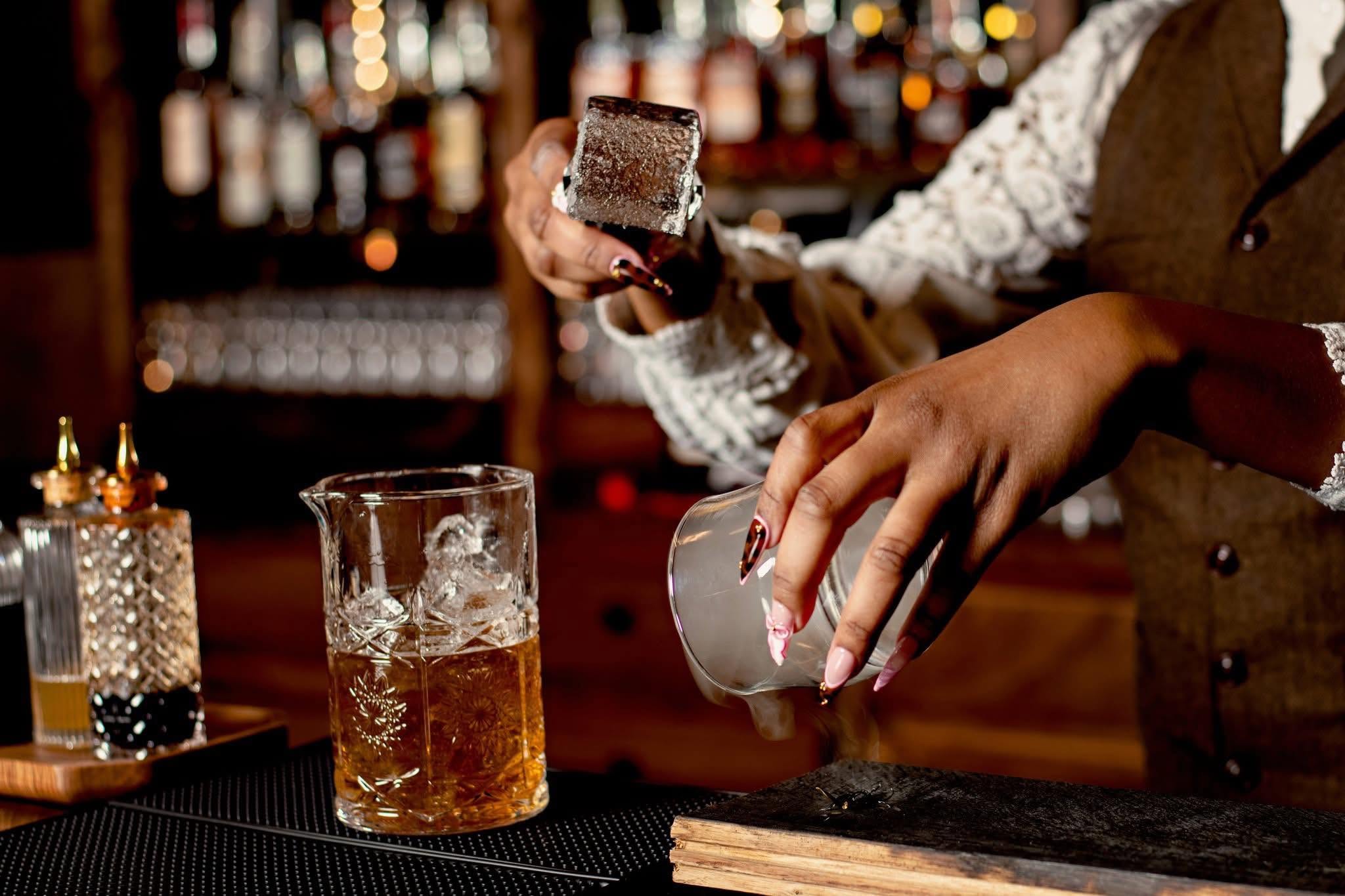 A bartender’s hands holding a large clear ice cube over a mixing glass, with a smoked rocks glass and bitters bottles on the bar