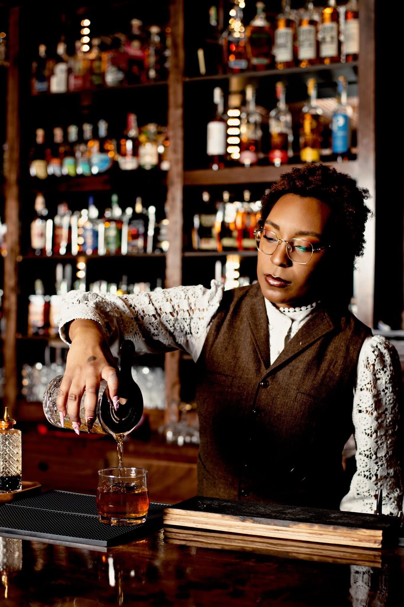 A bartender in a tweed vest straining a cocktail into a rocks glass, the back bar behind