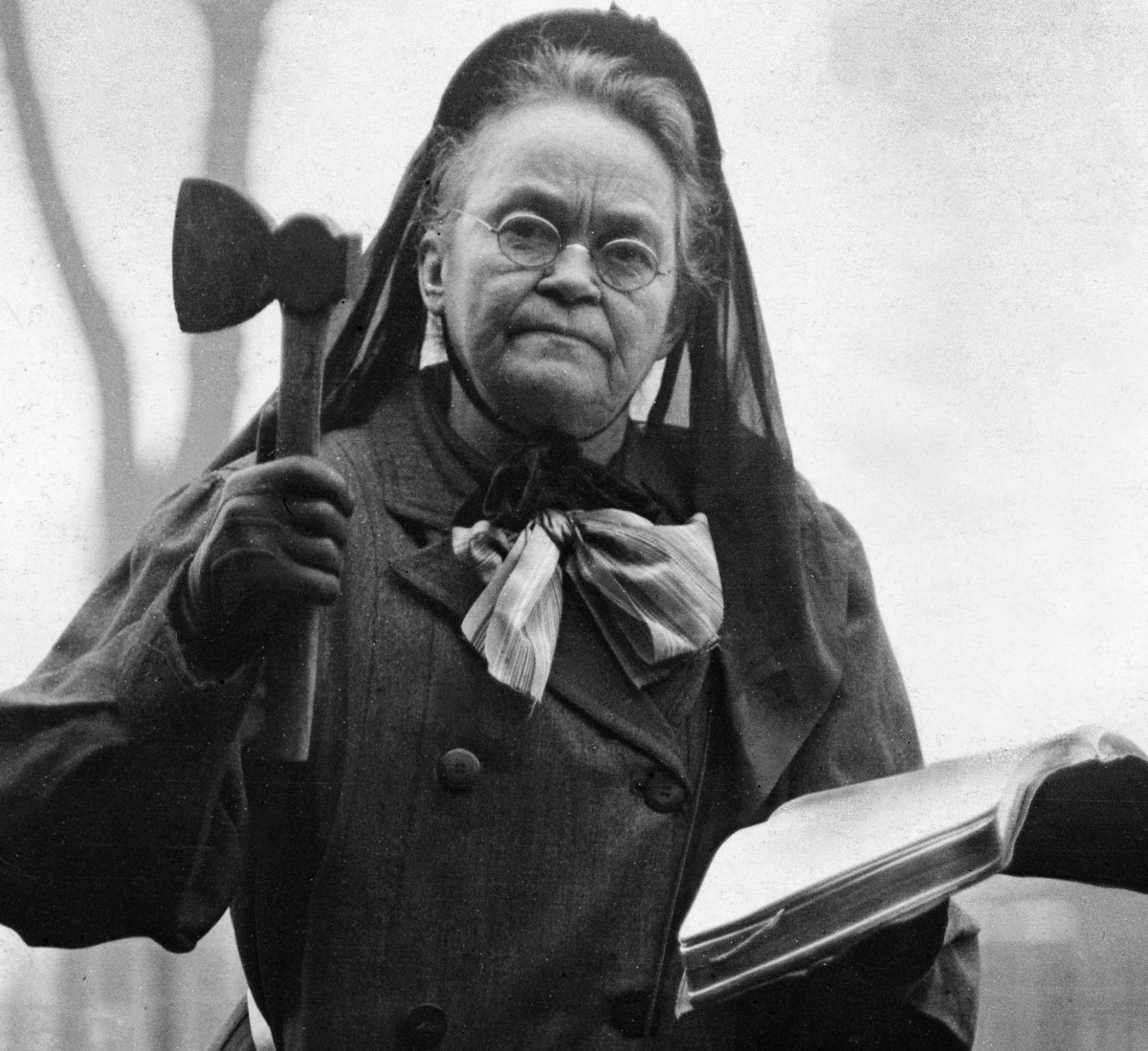 Carrie Nation in a dark coat and bonnet, holding a hatchet in one hand and an open Bible in the other